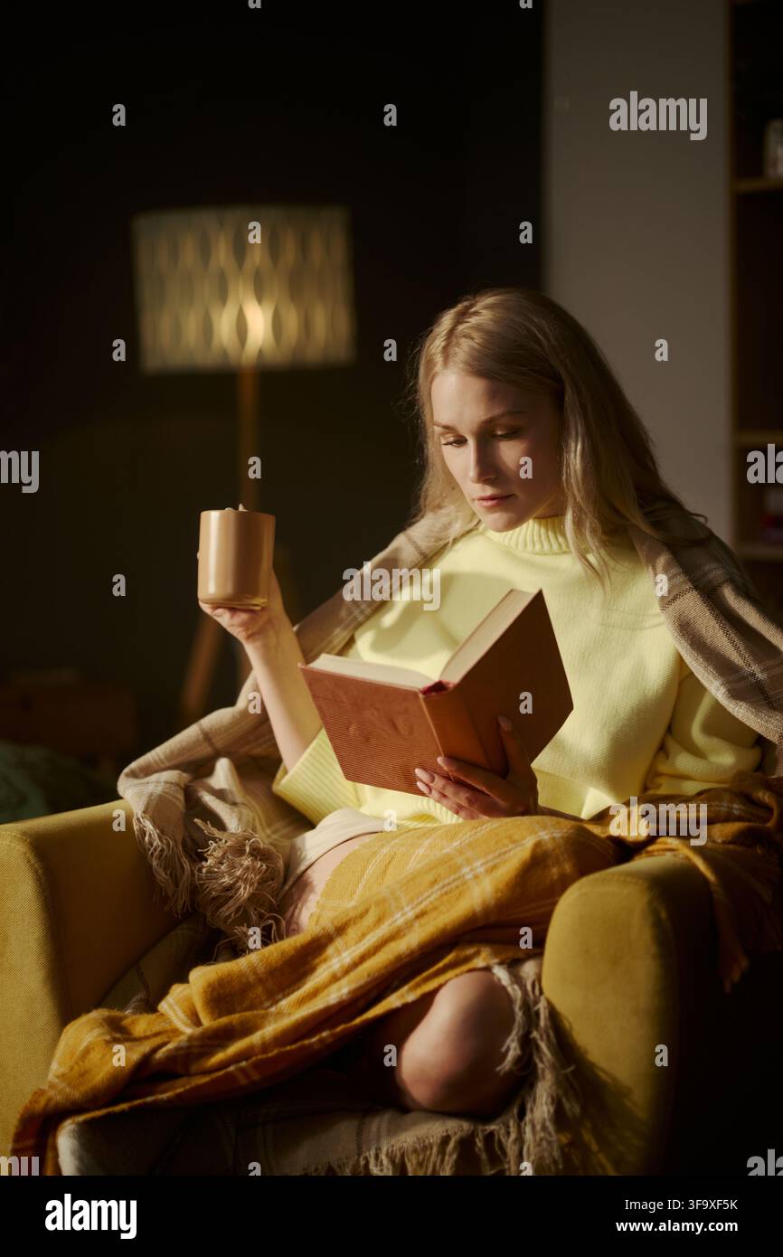 Giovane donna caucasica seduta su una poltrona, legge un libro e tiene una tazza. L'immagine rappresenta il benessere mentale e la cura di sé, adatta al marketing dello stile di vita e del tempo libero, verticale Foto Stock