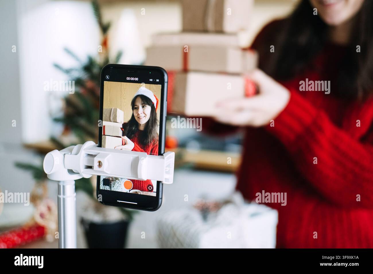 Telefono su treppiede filma donna con cappello da Babbo Natale che tiene una pila di pacchi incartati a casa. Marketing degli influencer per le festività, promozione video per le piccole imprese Foto Stock