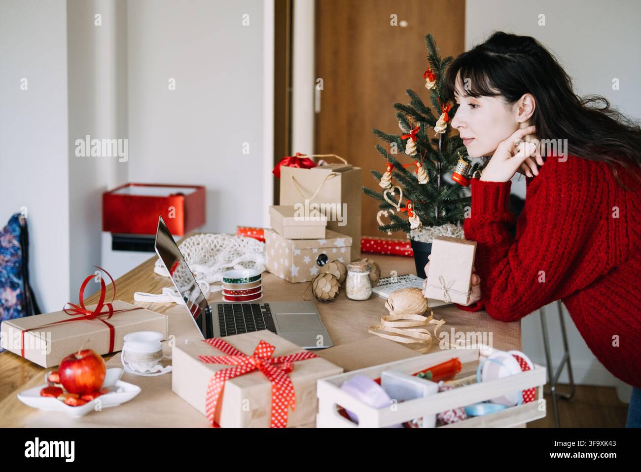 Donna si appoggia al tavolo tenendo un pacco kraft mentre guarda il laptop tra regali incartati. Ordini di Natale per piccole imprese, imballaggio di regali fatti a mano Foto Stock