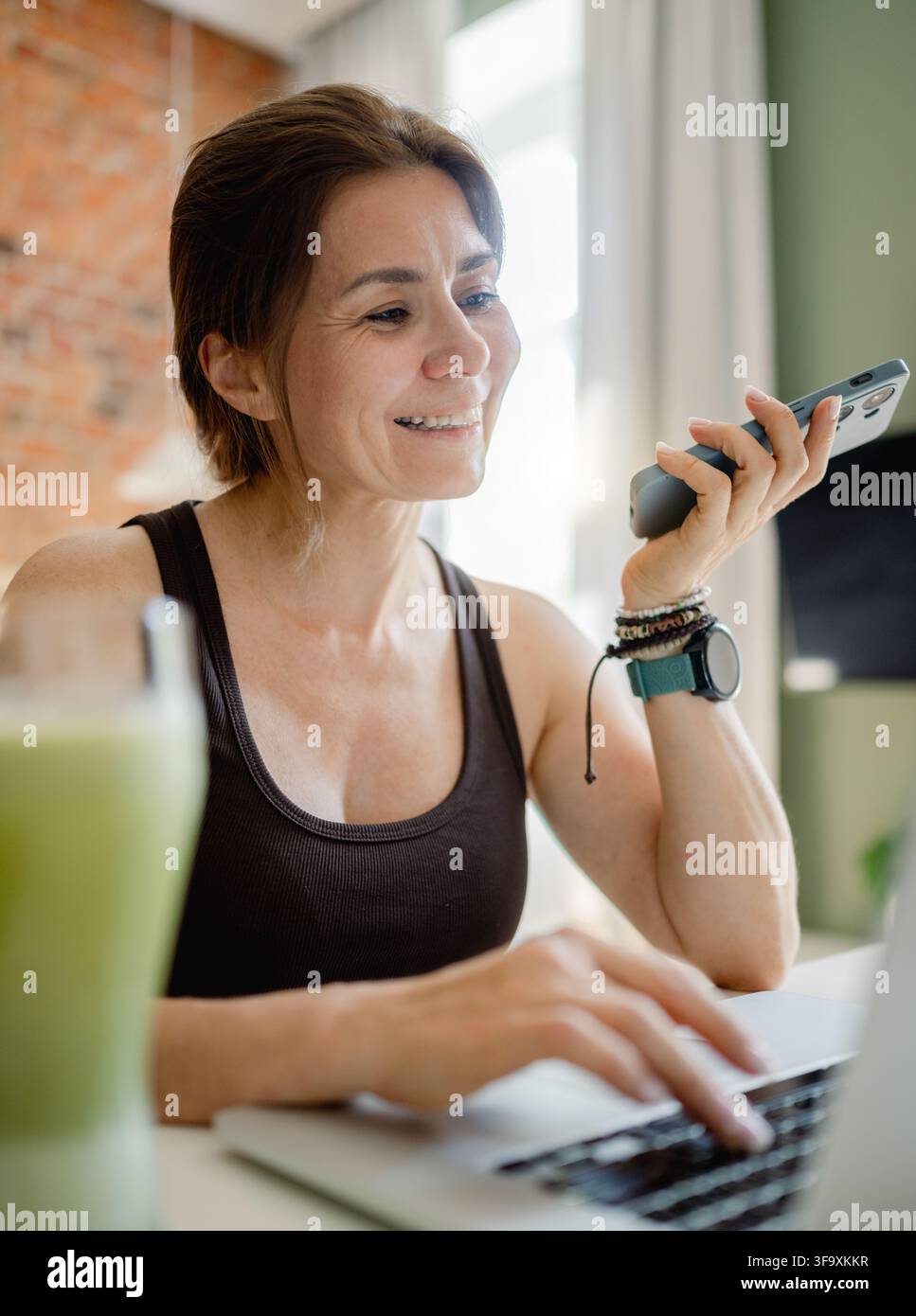 Donna sorridente lavora su laptop mentre parla al telefono nell'ufficio mattutino Foto Stock