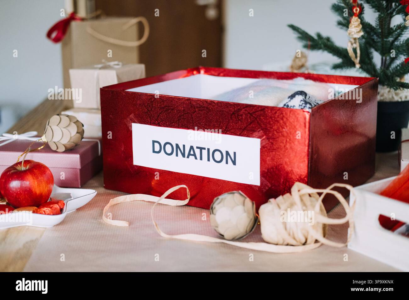 Scatola di DONAZIONE con maglieria piegata si trova tra regali incartati accanto all'albero di Natale. Raccolta fondi regali di Natale, donazione tra regali incartati Foto Stock