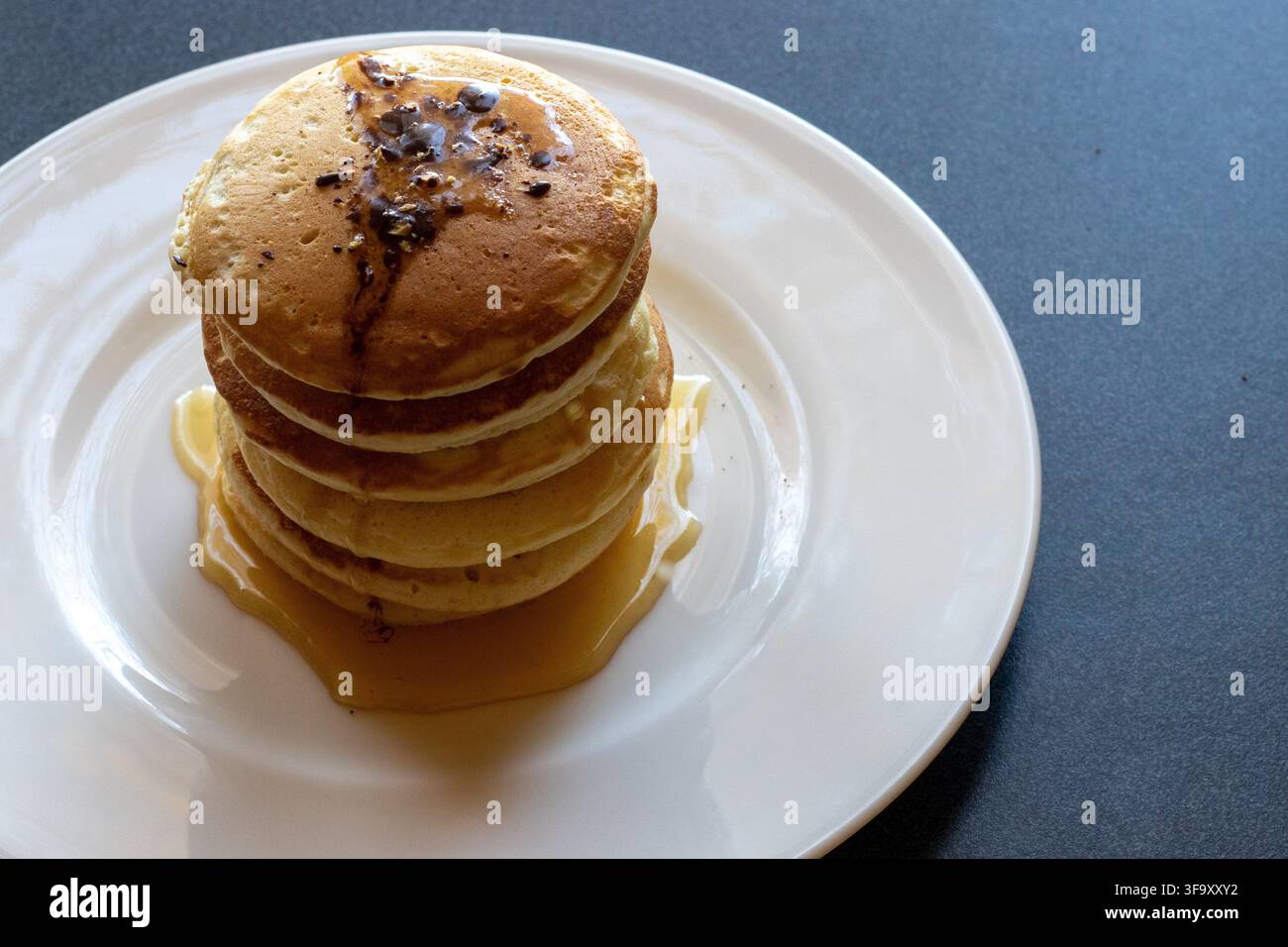 Vista dall'alto di una pila di pancake dorati glassati con cioccolato e sciroppo di miele su un piatto bianco, su uno sfondo scuro e suggestivo con spazio per il testo Foto Stock