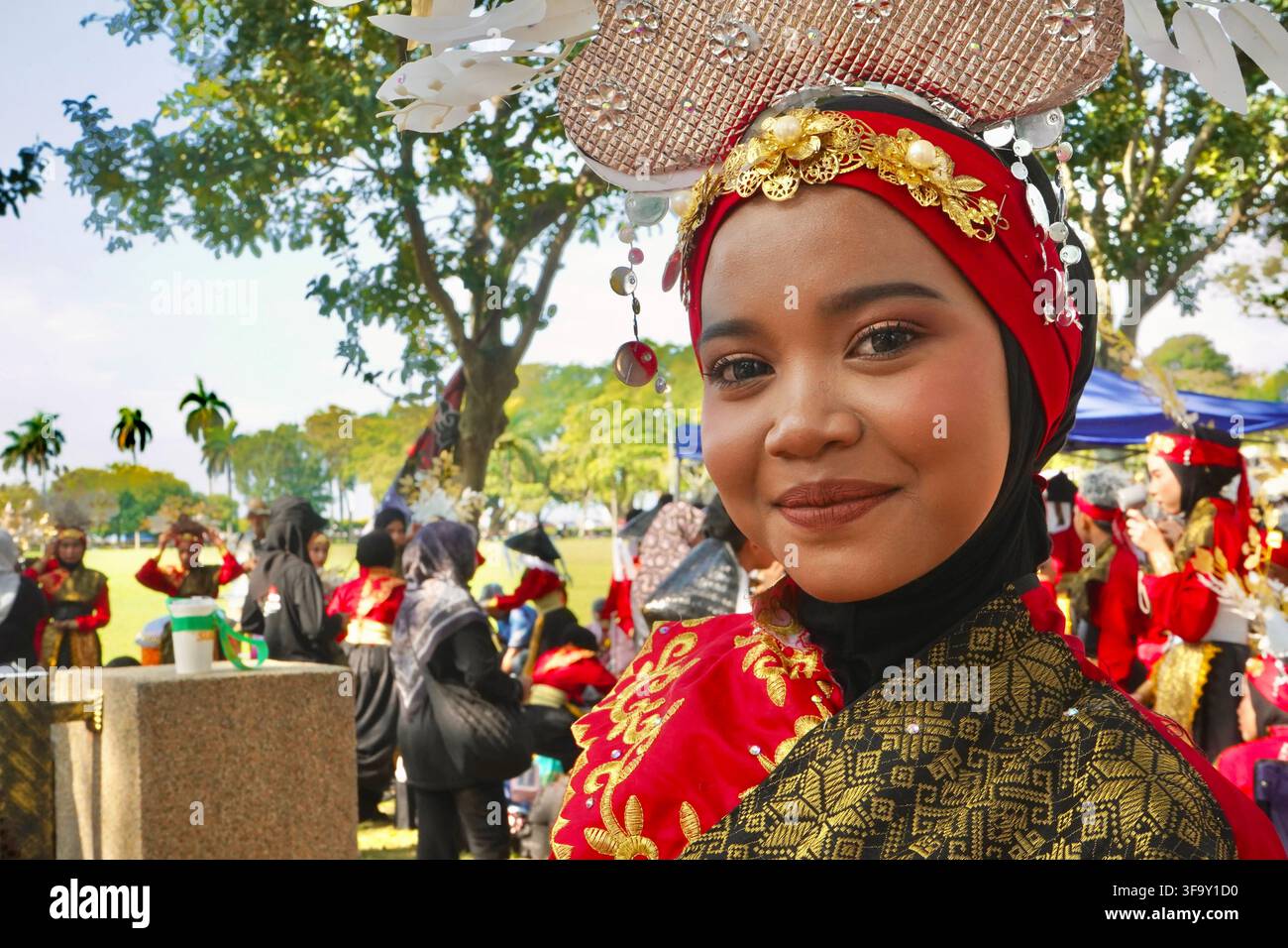 Giovane donna malese sorridente in costume tradizionale rosso e nero con copricapo dorato ornato durante un festival culturale all'aperto a George Town Penang, Malesia Foto Stock