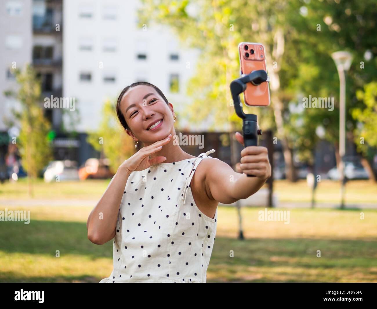 Creatrice di contenuti sorridente che si filma con uno stabilizzatore per smartphone durante una giornata soleggiata nel parco. Foto Stock