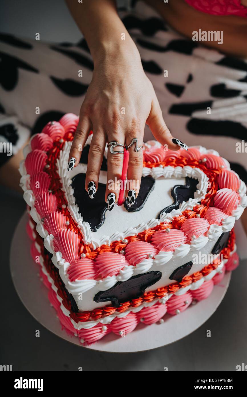 Mano che tocca una torta di compleanno a forma di cuore con glassa rosa e unghie a stampa mucca per la celebrazione Foto Stock