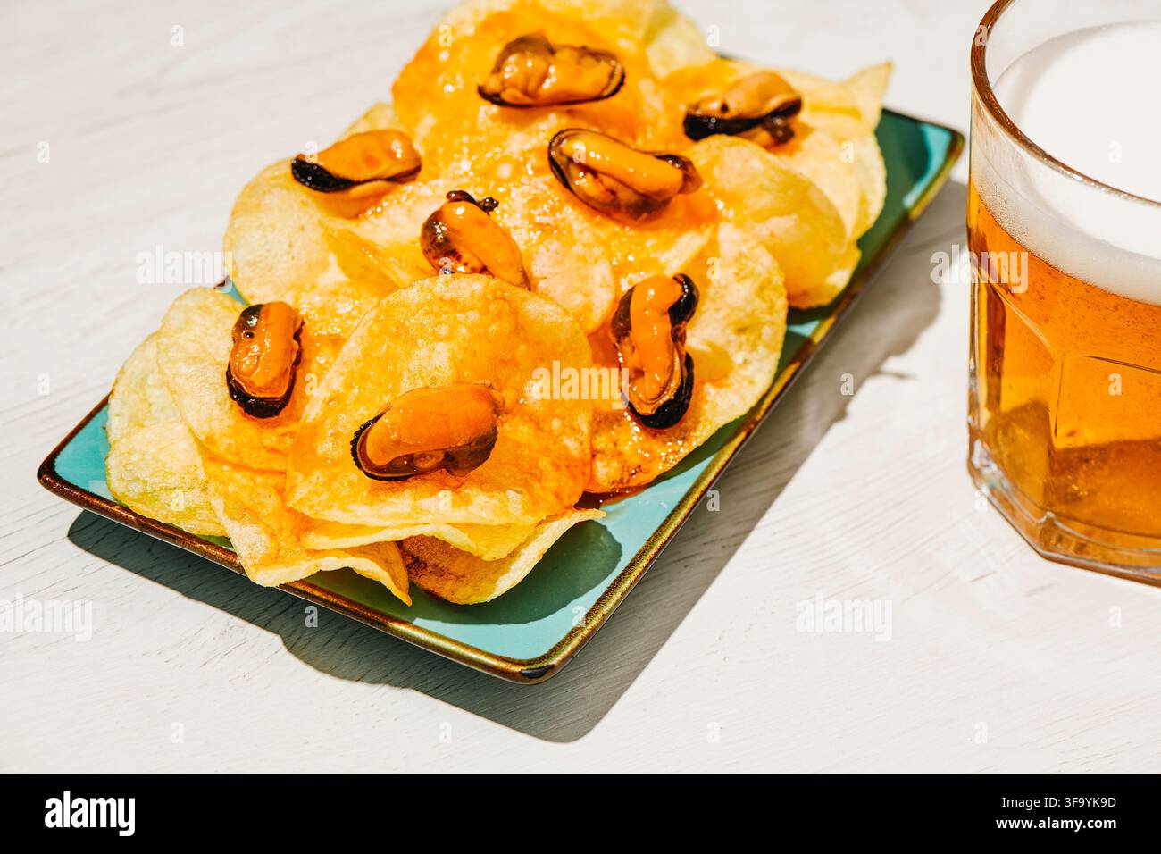 Un piatto di patatine fritte ricoperte di cozze sott'aceto è posato accanto a un bicchiere di birra su un tavolo di legno chiaro, una combinazione classica che si trova nei bar di tutta Sp Foto Stock