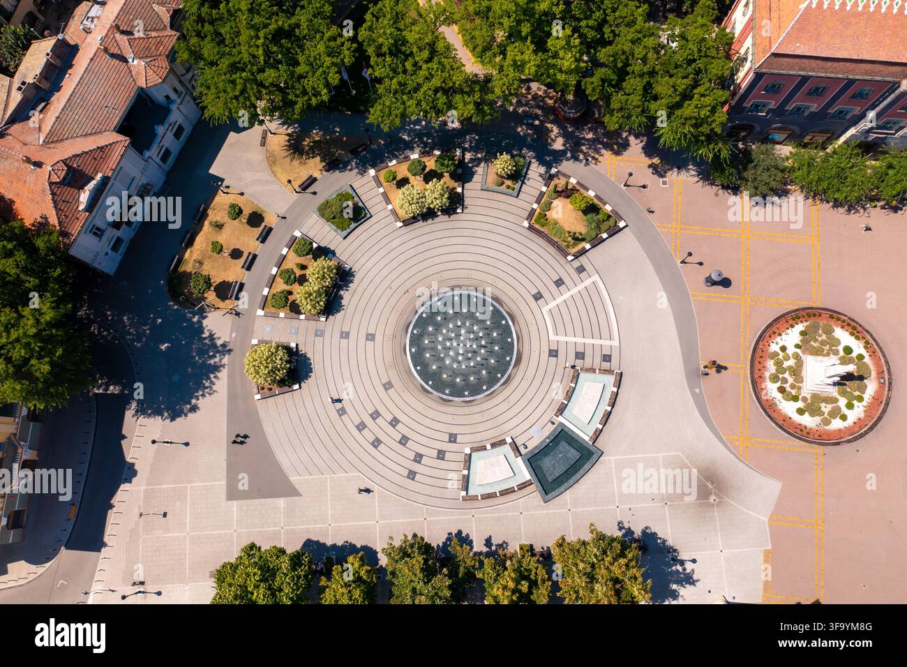 Vista aerea con drone di una fontana nella piazza principale della città di Subotica, in Serbia, circondata da edifici storici, spazi pubblici lastricati e verde urbano. Foto Stock
