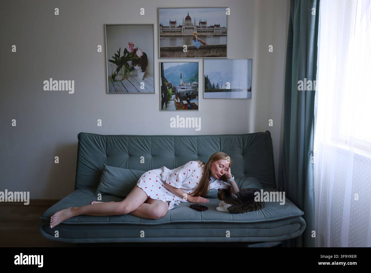 Donna sdraiata sul divano che utilizza lo smartphone mentre passa il tempo con il suo gatto in un soggiorno accogliente. Foto Stock