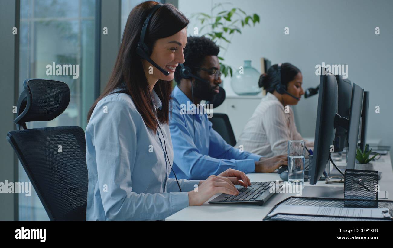 Donna d'affari caucasica sorridente che lavora in un call center con una squadra multiculturale di colleghi operatori che parlano su una linea di assistenza con auricolare e computer Foto Stock