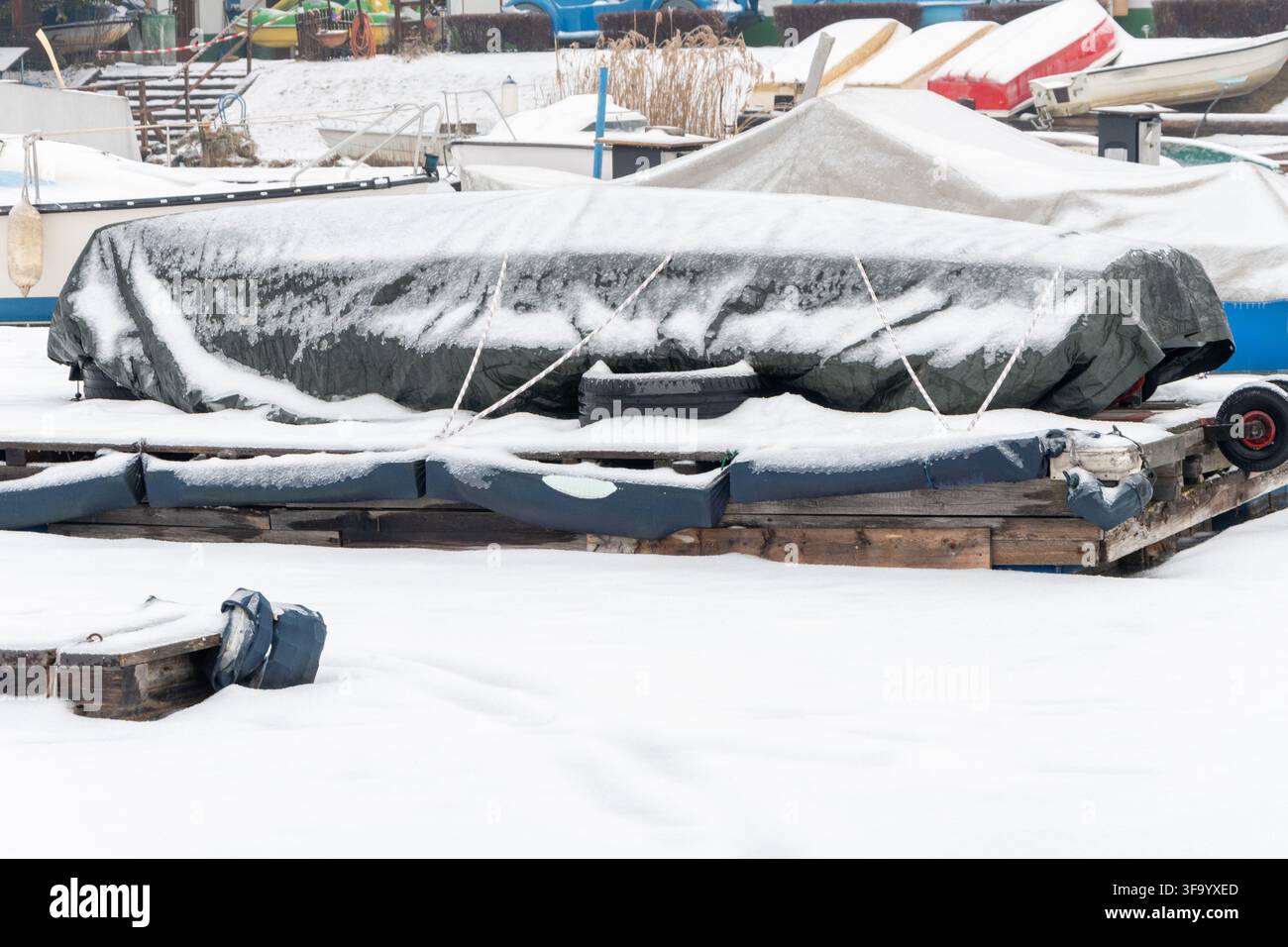 Una barca coperta da un telone è ormeggiata su un molo innevato tra altre piccole barche in una marina invernale. Foto Stock