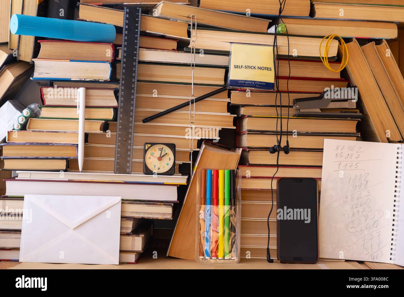 Uno spazio di lavoro ingombro creato da libri impilati contiene penne, pennarelli colorati, righello, orologio, post-it, smartphone, auricolari e un blocco note. Foto Stock