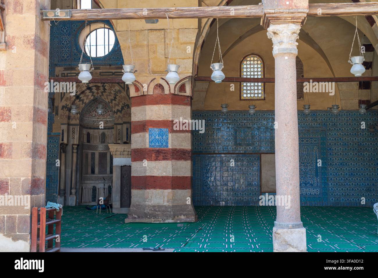 Vista dell'interno della storica moschea di Aqsunqur con piastrelle blu di Iznik, colonne in pietra a strisce, lanterne sospese e tappeto da preghiera verde a E Foto Stock