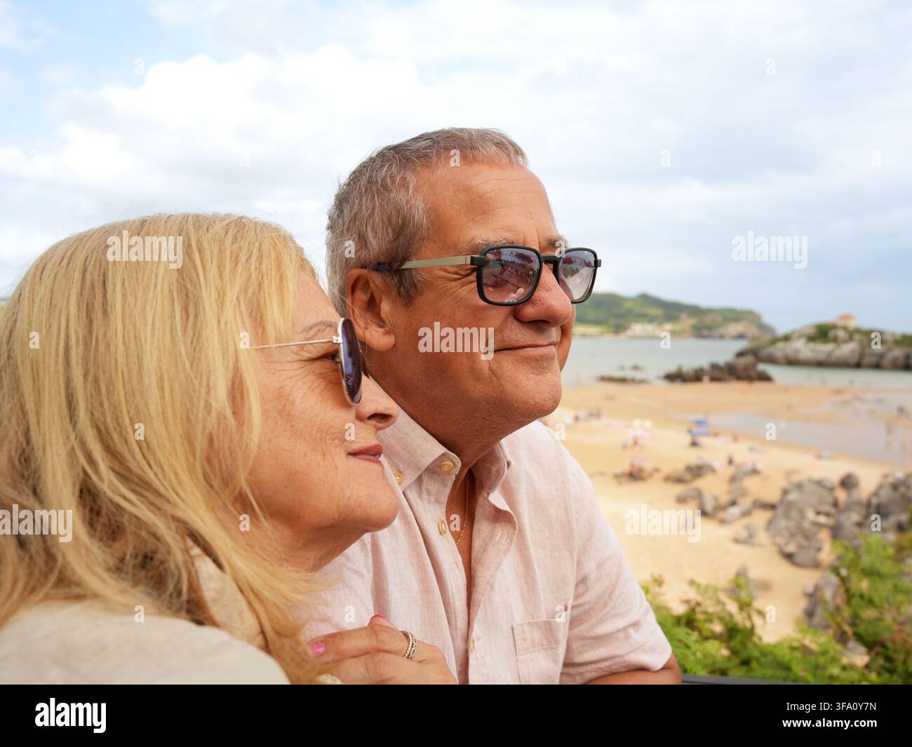Coppia di anziani sorridenti con occhiali da sole si rilassa insieme sopra una spiaggia sabbiosa, godendosi una tranquilla vacanza costiera con vista sul mare, luce calda del giorno e l Foto Stock
