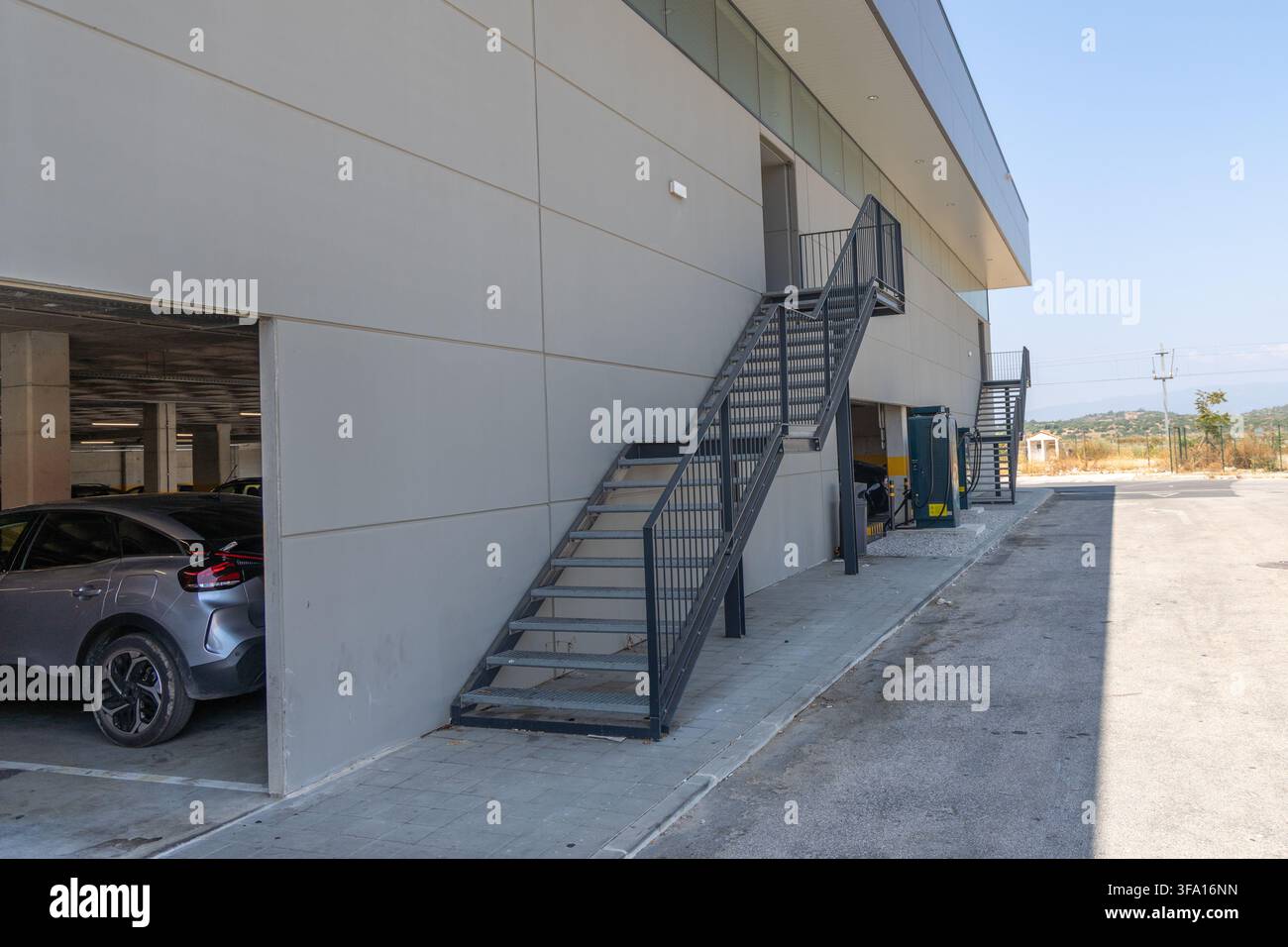 Una vista esterna di una struttura commerciale contemporanea con scale metalliche esterne e un'anteprima di un parcheggio con un veicolo Foto Stock