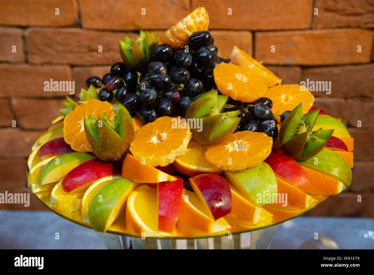 Vassoio di frutta fresca splendidamente disposto con mele, arance, kiwi e uva nera su uno sfondo di muro di mattoni. Catering gourmet salutare Foto Stock