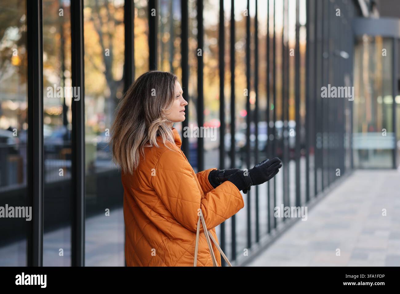 donna pensierosa in giacca trapuntata arancione e guanti neri che fa shopping in una strada urbana con vetrine in vetro periodo periodo periodo periodo periodo periodo Foto Stock