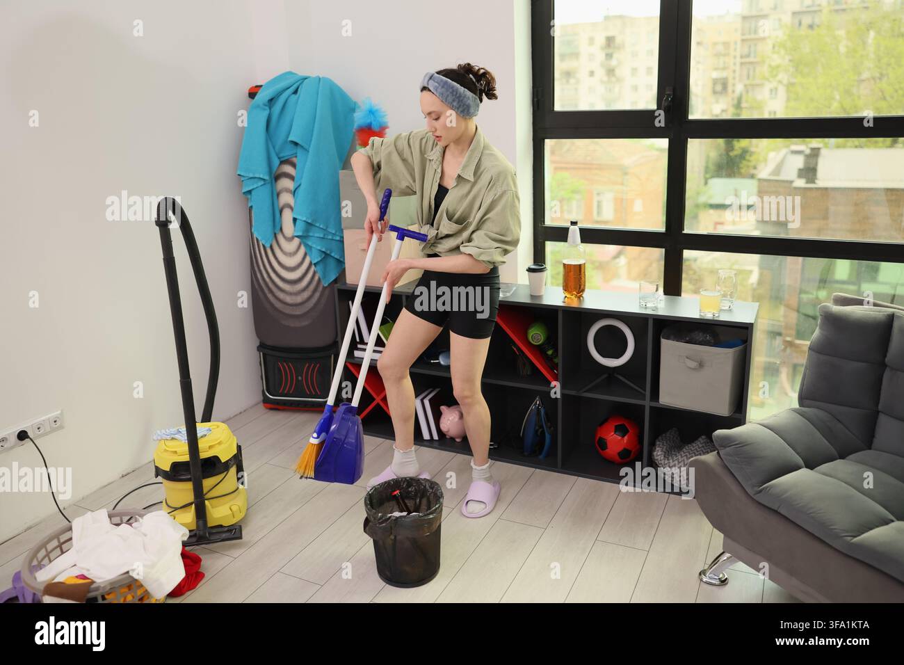 Giovane donna che pulisce un appartamento moderno con mocio e paletta accanto a uno scaffale di stoccaggio, aspirapolvere, cesto della biancheria e una grande finestra sulla città, mostrando la vita domestica Foto Stock