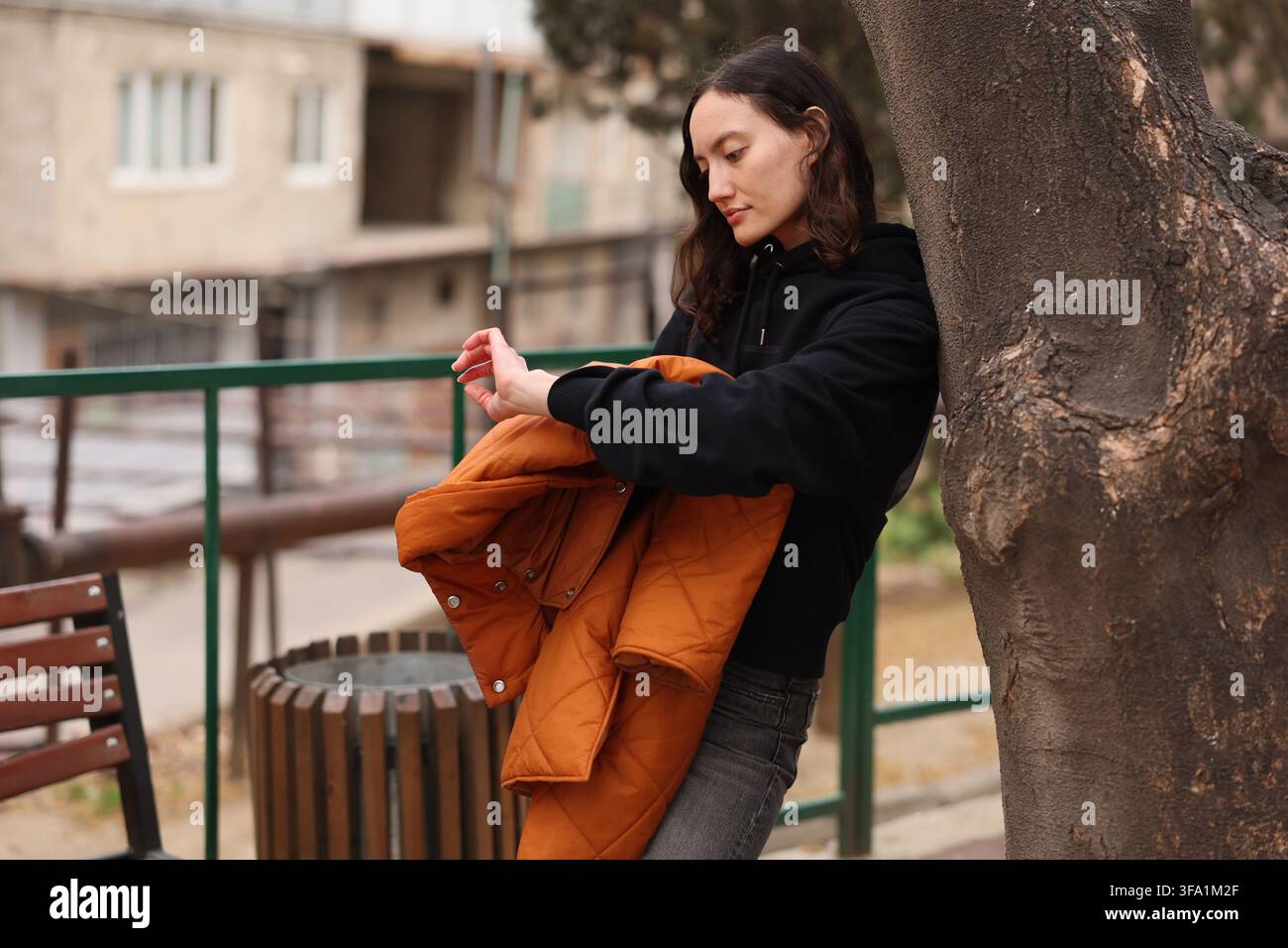 Giovane donna pensierosa in felpa con cappuccio nera che tiene una giacca arancione mentre si appoggia a un albero in un tranquillo cortile urbano con edifici sfocati in morbido, parco Foto Stock