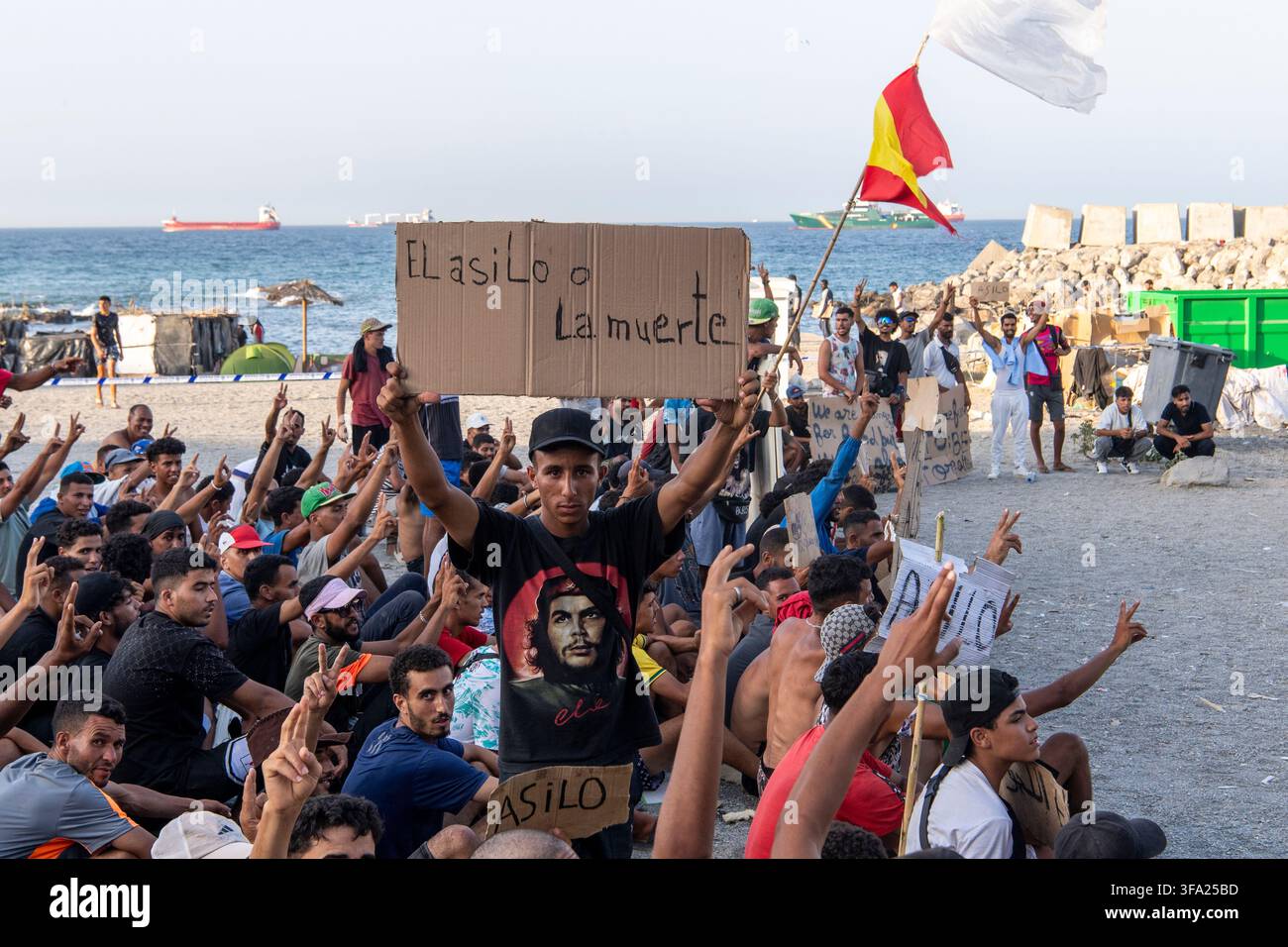 CEUTA, SPAGNA - 17 AGOSTO 2026: Più di 100 migranti, la maggior parte dei quali marocchini, protestano da giorni davanti ai furgoni della Polizia Nazionale durante la distribuzione giornaliera di cibo fornita dalla Croce Rossa spagnola alla spiaggia Trampolín. “Nessun asilo, nessun cibo” è il canto ripetuto dai migranti sulla sabbia, mentre i migranti africani continuano a rimanere nelle strutture di alloggio temporanee allestite sulla spiaggia di Ceuta. Le autorità dovrebbero sgomberare la spiaggia nei prossimi giorni e trasferire i migranti nei quattro centri di accoglienza annunciati oggi dal Ministro spagnolo per la Migrazione, Elma Saiz. Foto di Foto Stock