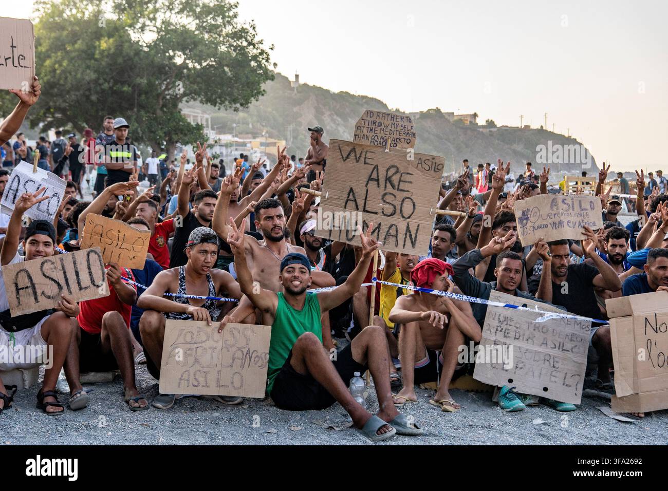CEUTA, SPAGNA - 17 AGOSTO 2026: Più di 100 migranti, la maggior parte dei quali marocchini, protestano da giorni davanti ai furgoni della Polizia Nazionale durante la distribuzione giornaliera di cibo fornita dalla Croce Rossa spagnola alla spiaggia Trampolín. “Nessun asilo, nessun cibo” è il canto ripetuto dai migranti sulla sabbia, mentre i migranti africani continuano a rimanere nelle strutture di alloggio temporanee allestite sulla spiaggia di Ceuta. Le autorità dovrebbero sgomberare la spiaggia nei prossimi giorni e trasferire i migranti nei quattro centri di accoglienza annunciati oggi dal Ministro spagnolo per la Migrazione, Elma Saiz. Foto di Foto Stock