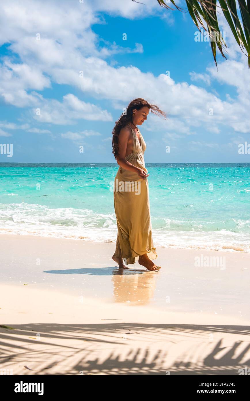 Una donna con un vestito maxi beige passeggia lungo la costa tropicale sabbiosa dell'isola di Dhigurah alle Maldive accanto alle onde turchesi dell'oceano sotto un sole Foto Stock
