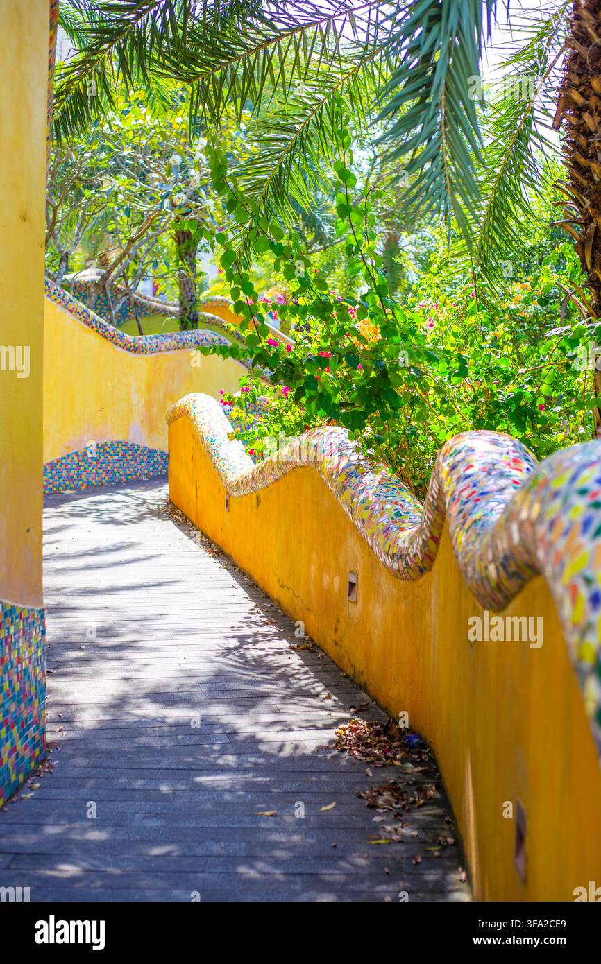 Un sentiero tortuoso in un parco mediterraneo con pareti a mosaico giallo ondulate e palme in prospettiva verticale. Progettazione creativa del paesaggio del parco. Foto Stock