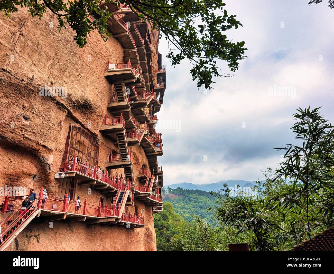 Vista delle grotte storiche di Maijishan con intricati passaggi in legno intagliati nella ripida parete di scogliera di arenaria rossa con una lussureggiante foresta verde a T Foto Stock