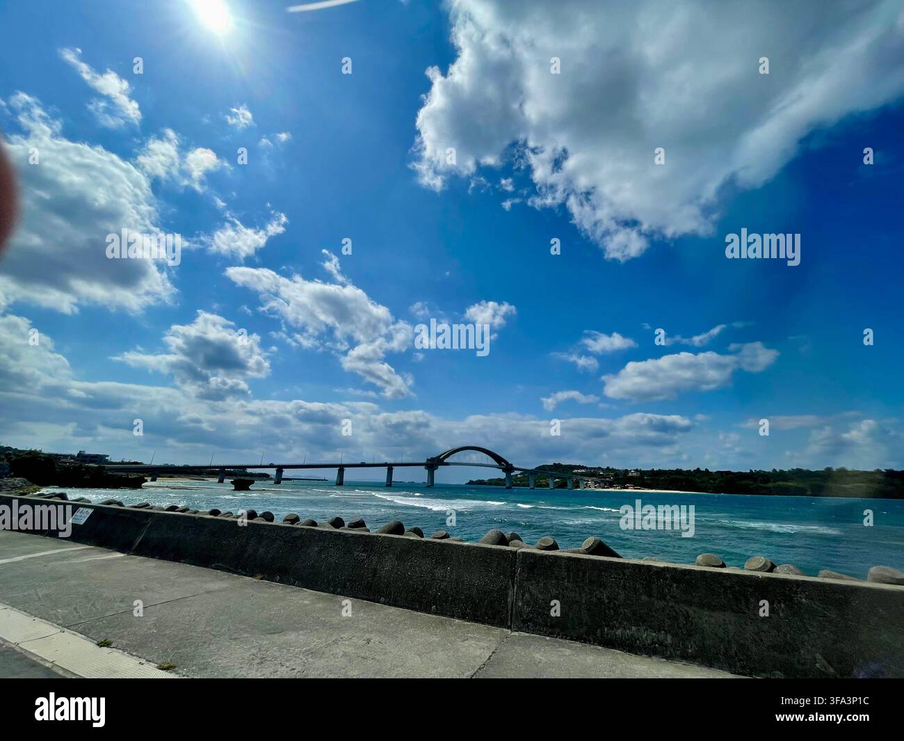 Una vista ampia di un moderno ponte ad arco che si estende su acque costiere blu sotto un cielo luminoso con nuvole bianche sparse. okinawa giappone - Immagine stock catturata con smartphone