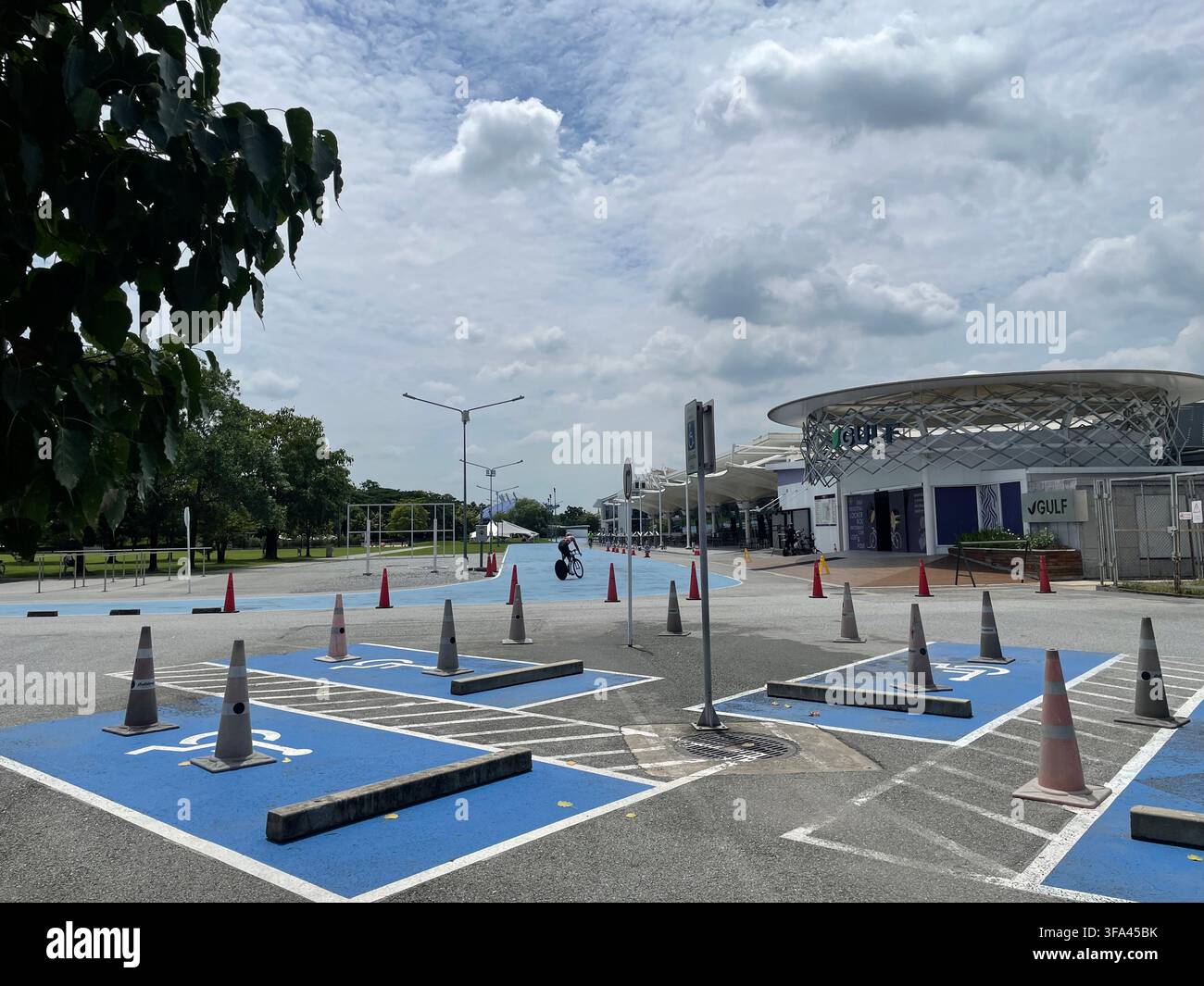 Una vista di un parcheggio accessibile dipinto di blu davanti a un edificio moderno con tetto circolare sotto un cielo parzialmente nuvoloso, con più coni di segnalazione. - Immagine stock catturata con smartphone