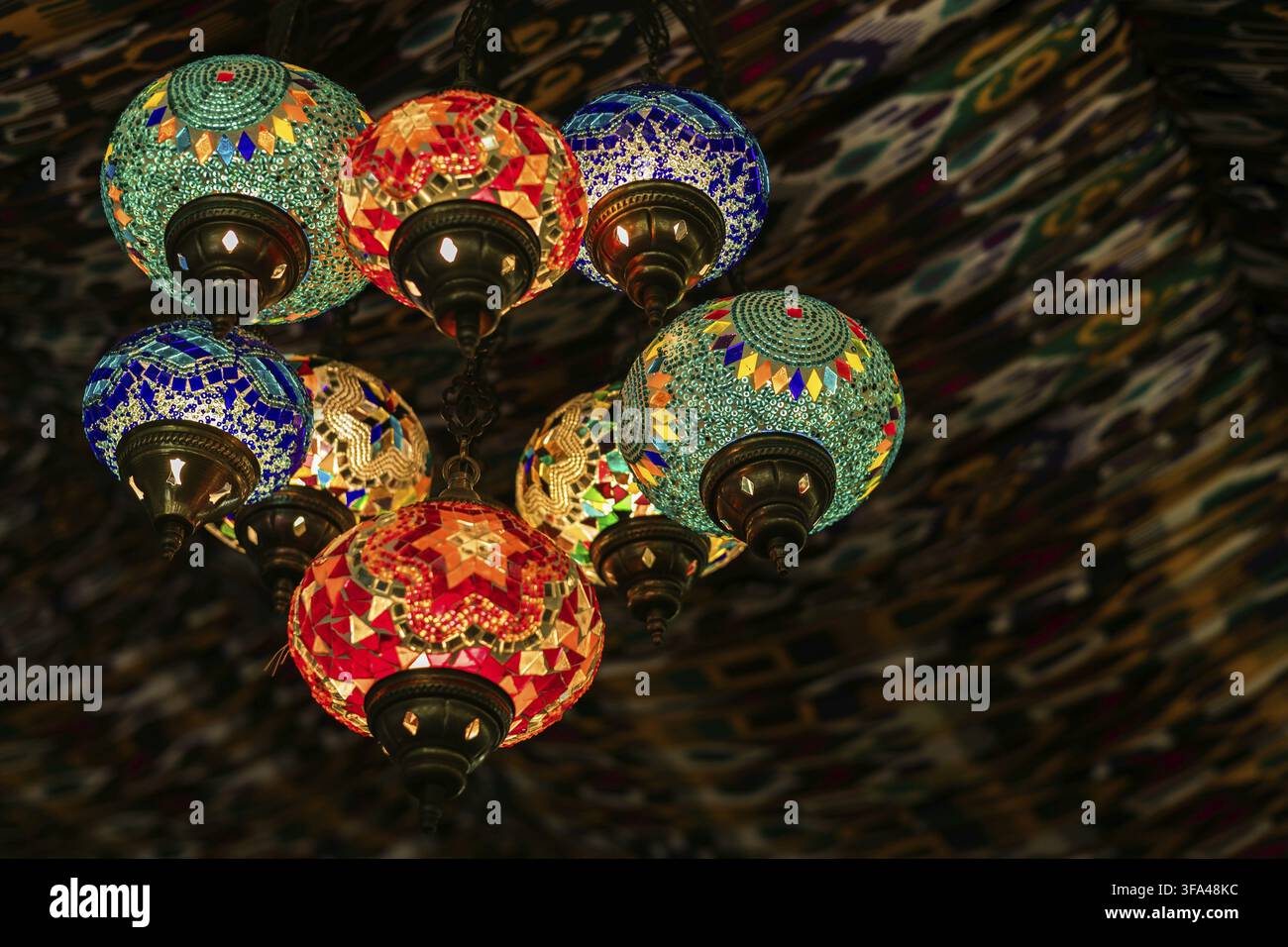 Gruppo ornato di lampade a mosaico marocchine o turche luminose con motivi geometrici colorati appese da un soffitto decorativo, creando un'atmosfera calda ed esotica Foto Stock
