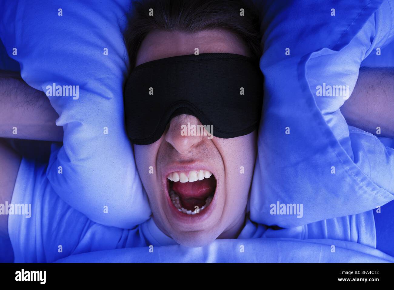 Un uomo arrabbiato che indossa una maschera per dormire urla nel letto mentre si copre le orecchie con i cuscini. L'immagine rappresenta l'insonnia, la mancanza di sonno o un nitrito rumoroso Foto Stock