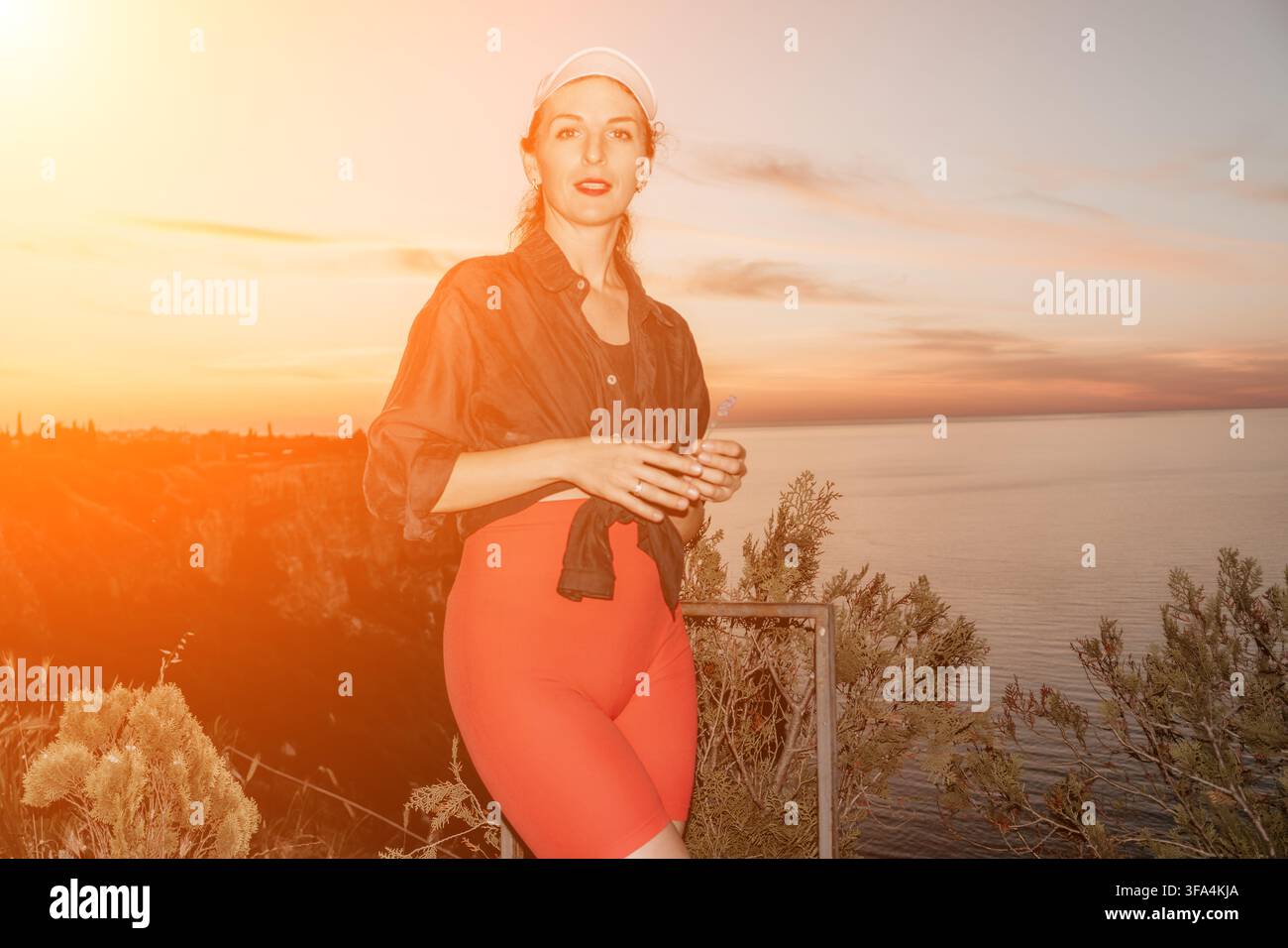 Sfondo vivace del tramonto incornicia una giovane donna elegante con cappellino e abiti casual in piedi all'aperto vicino alle scogliere costiere Foto Stock