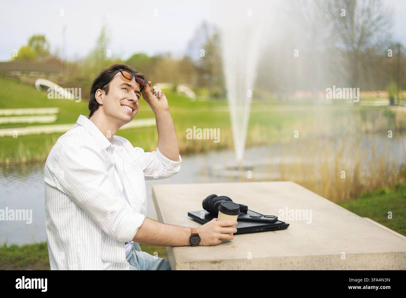 Un uomo sorridente che lavora sul suo laptop in un parco illuminato dal sole accanto a una fontana. Un concetto per il freelance, lo stile di vita dei digital nomad e la flessibilità Foto Stock