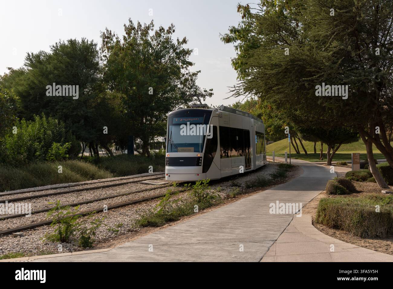 Education City, Al Rayyan, Qatar – 4 febbraio 2026: Una vista frontale del moderno tram bianco con linea gialla che arriva sui binari attraverso un parco verde. Foto Stock