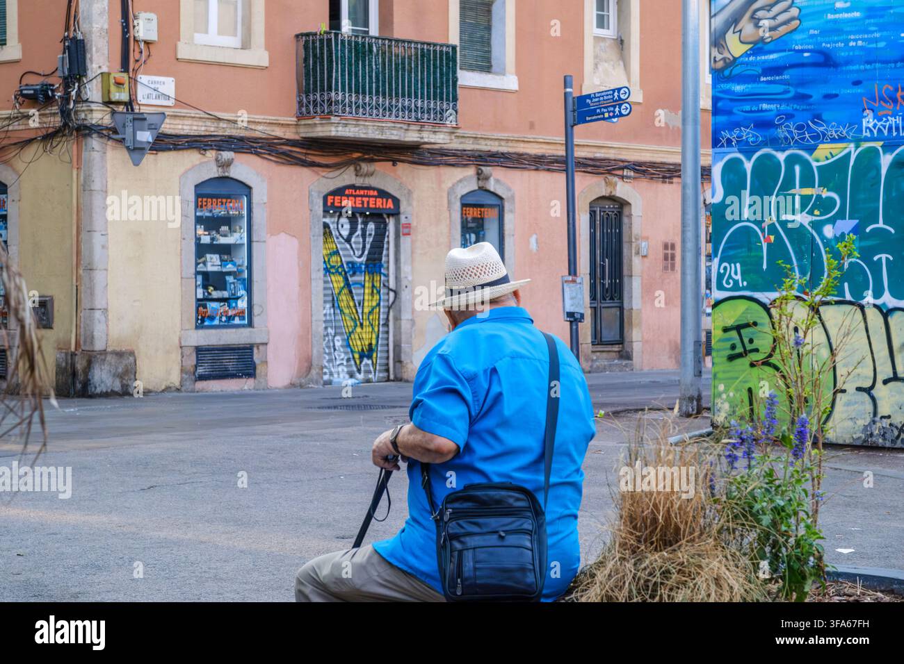 Barcellona – Spagna – 1 agosto 2026: Anziano seduto in strada nel quartiere di Barceloneta, camicia blu e cappello di paglia, vista posteriore, negozio tradizionale Foto Stock