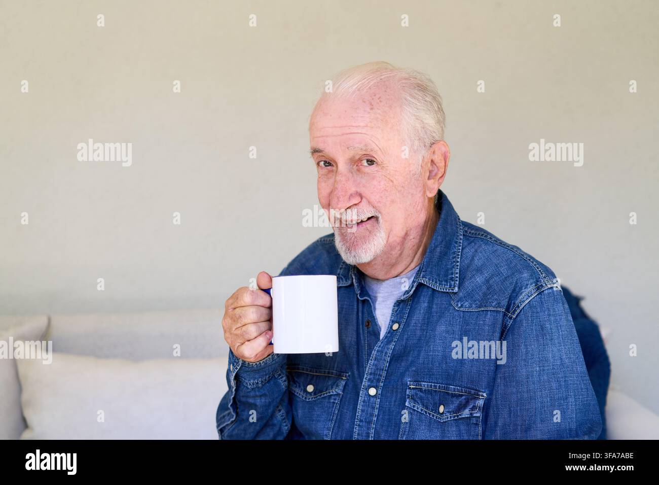 Invecchiamento sano e vita indipendente, uomo anziano che gode di una bevanda calda a casa sua. Paziente maschio anziano in un soggiorno luminoso e minimalista, che promuove Foto Stock