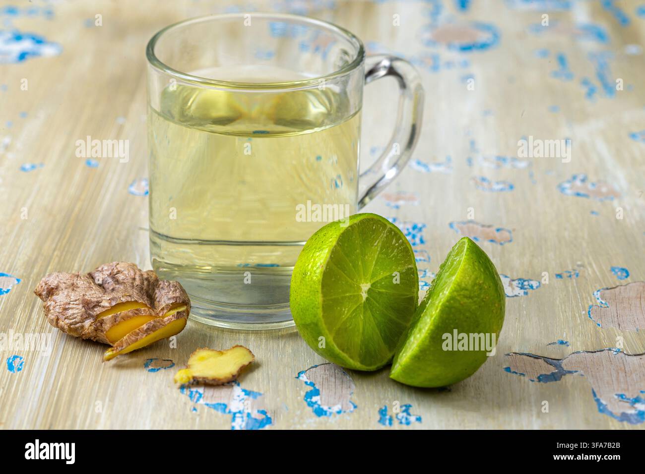 Una tazza di vetro contenente un liquido giallo, accompagnata da Zingiber officinale e Citrus aurantiifolia affettati su una superficie di legno consumata. Foto Stock