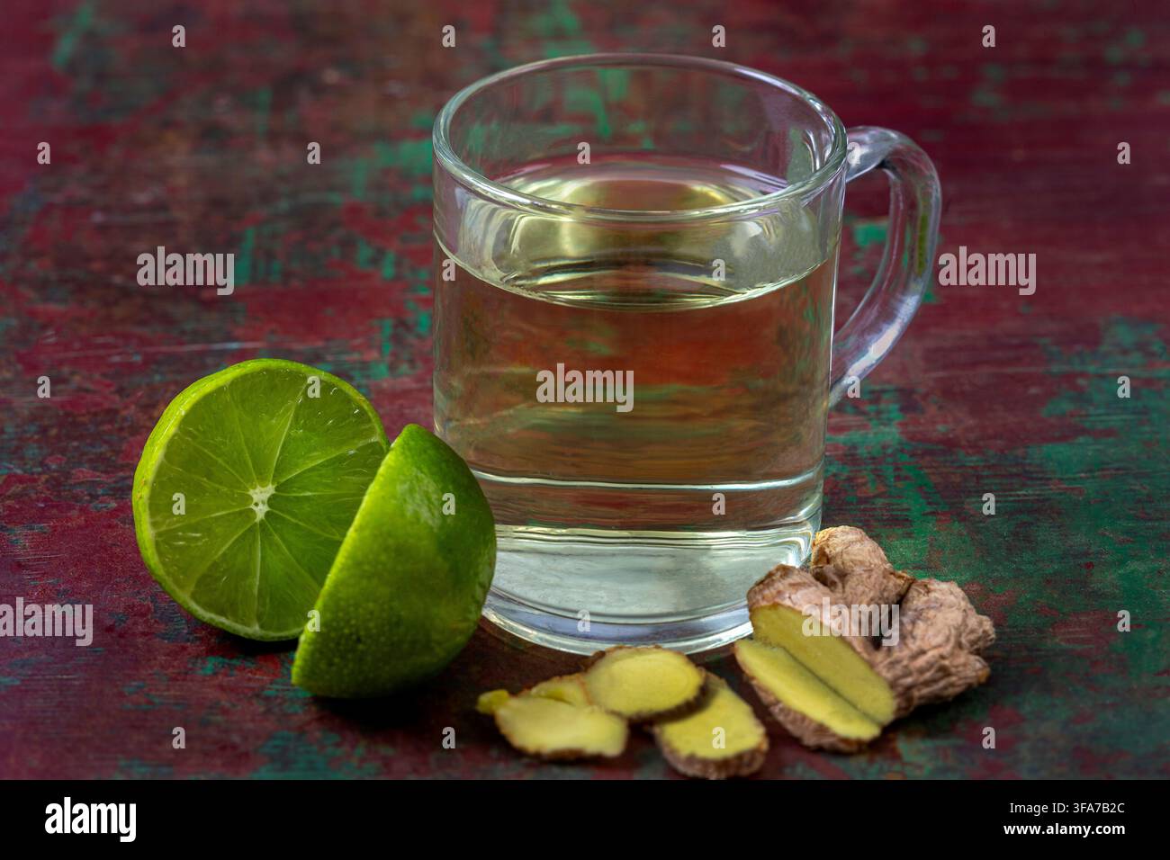 Una tazza di vetro contenente un liquido giallo pallido, accompagnata da Citrus aurantiifolia affettata e rizoma di Zingiber officinale affettato su una superficie strutturata. Foto Stock
