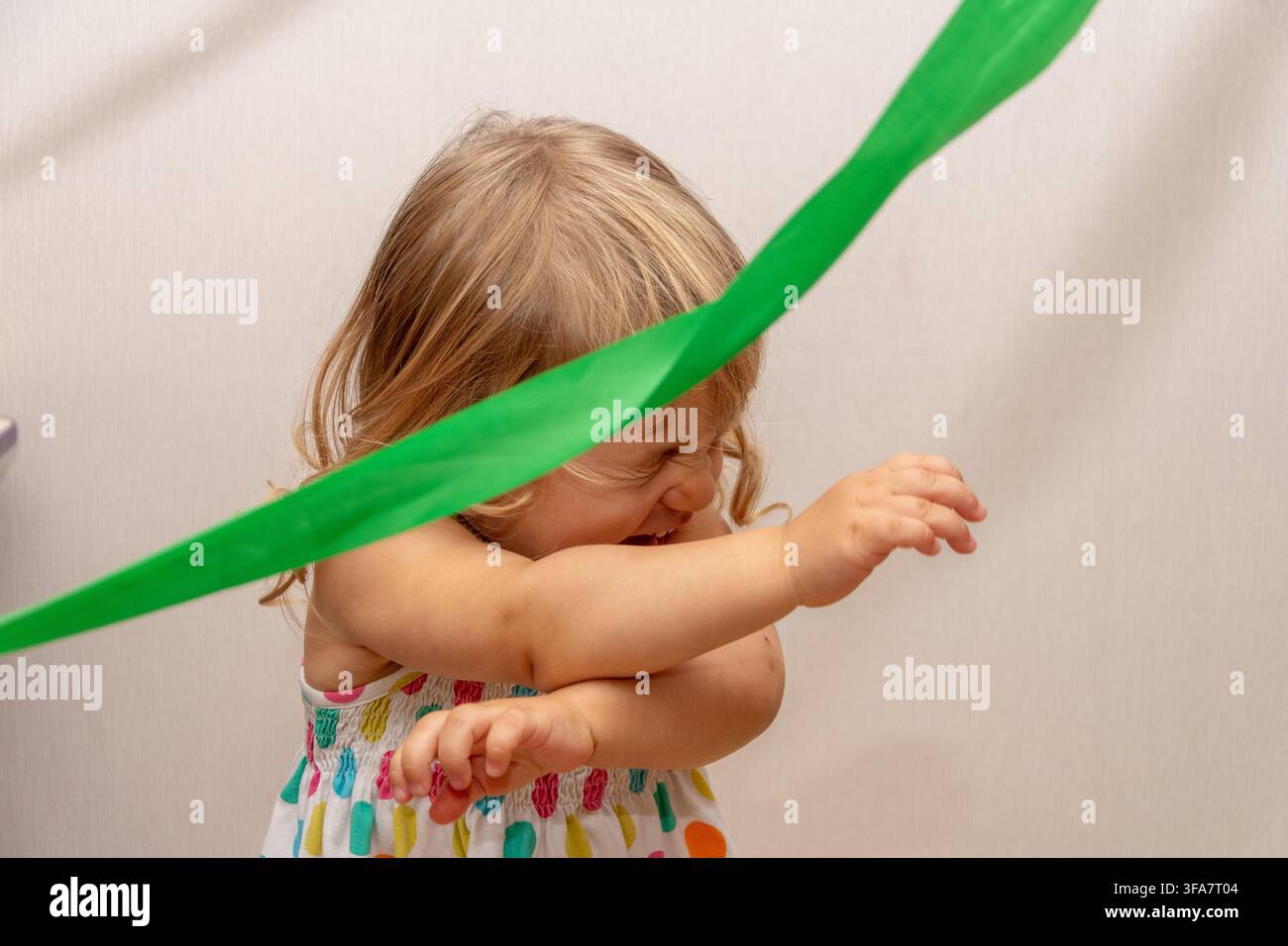 Una bambina che ride in un vestito colorato alza le braccia mentre un nastro verde le passa davanti al viso. Foto Stock