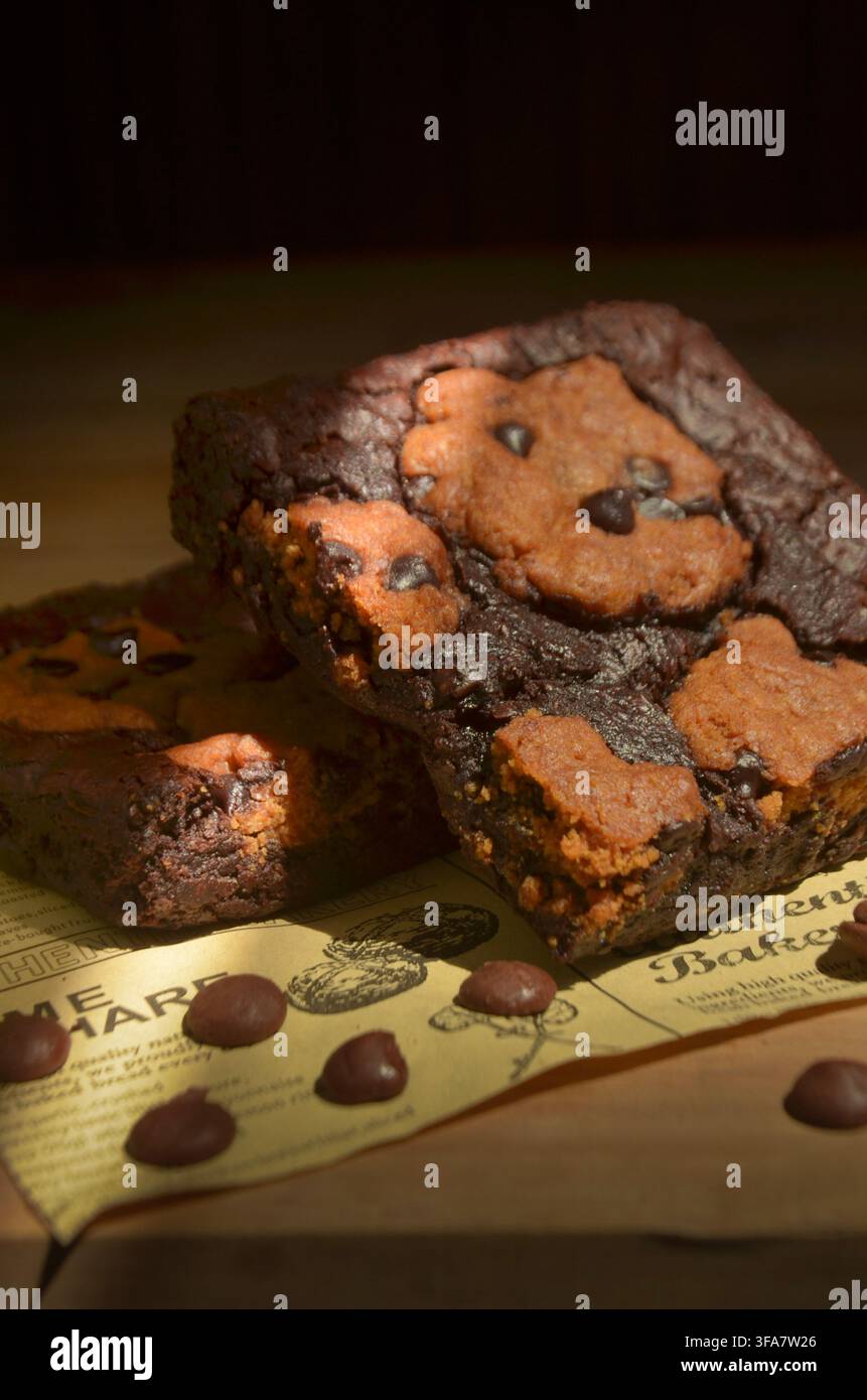 Brownies ricchi al cioccolato con gocce di cioccolato si adagiano su carta vintage. L'illuminazione calda mette in risalto la loro consistenza umida e densa. Ogni morso promette una profondità, deca Foto Stock