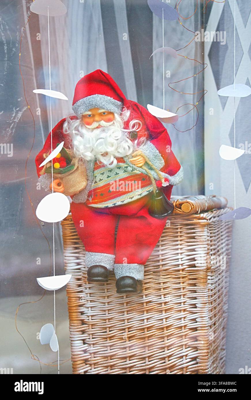 Una figura allegra di Babbo Natale seduta su un cesto di vimini in un interno accogliente adornato con decorazioni festive, caratterizzato da colori rossi e bianchi, un pulito Foto Stock