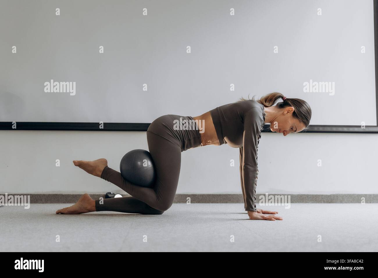 Donna che fa esercizio con il fitball al chiuso. Giovane donna mantiene una posa di equilibrio in ginocchio con una piccola palla da esercizio in uno studio domestico tranquillo, mostrando Foto Stock
