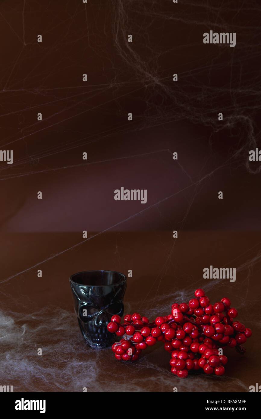 Una tazza di vetro nera a forma di teschio che si trova accanto a un grappolo di bacche rosse vivaci, posizionata su una superficie scura stratificata con delicate ragnatele, davanti a Foto Stock