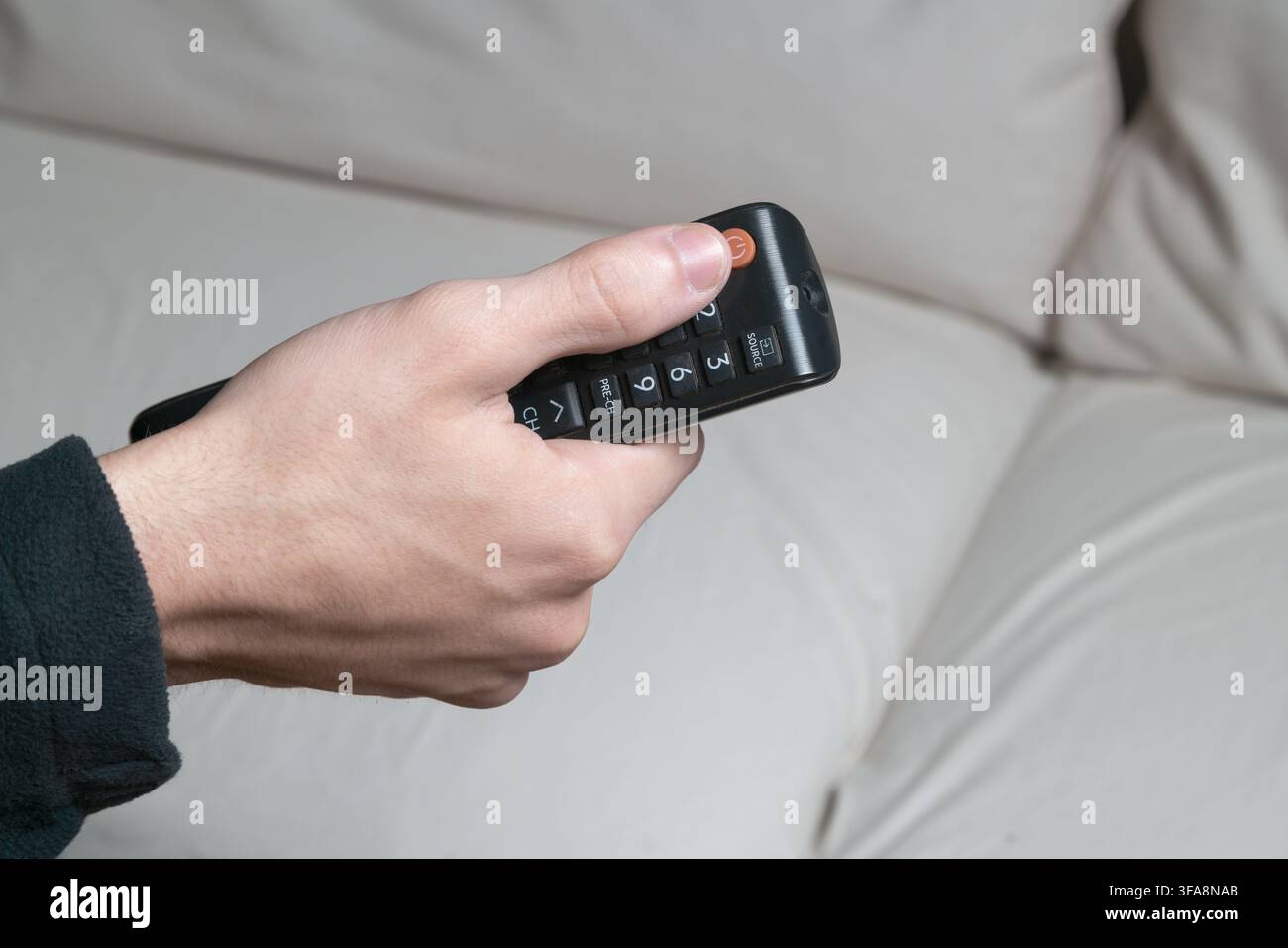 Una mano accende il telecomando TV con un divano bianco sullo sfondo Foto Stock