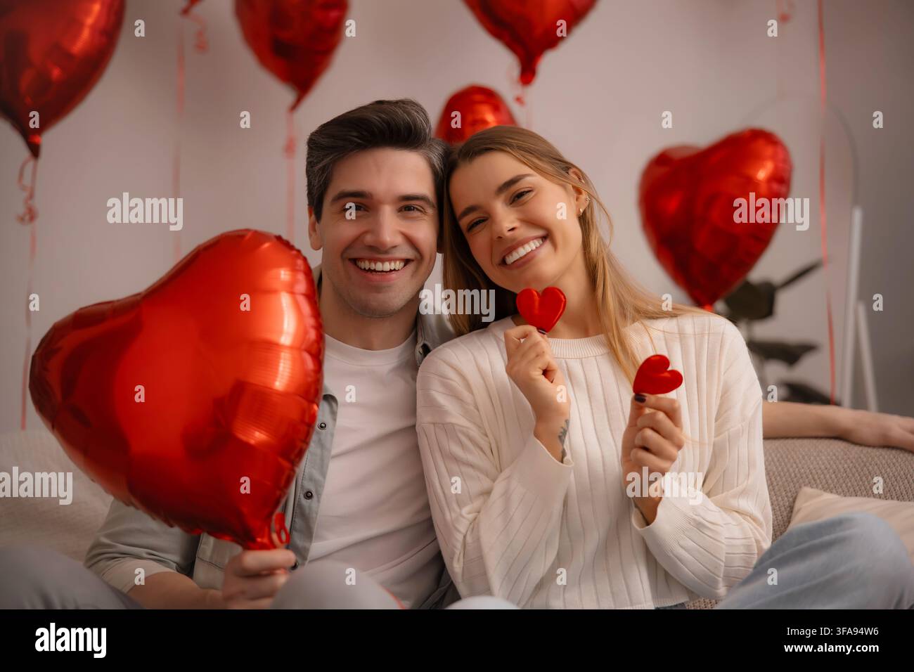 Una giovane coppia allegra seduta insieme su un divano con palloncini a forma di cuore rossi e piccole decorazioni a forma di cuore durante una celebrazione di San Valentino a casa. Foto Stock