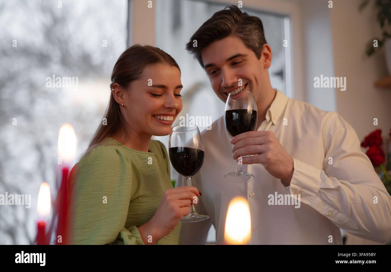 Giovane coppia sorridente solleva bicchieri di vino rosso accanto a candele calde durante una romantica celebrazione di San Valentino a casa. Foto Stock