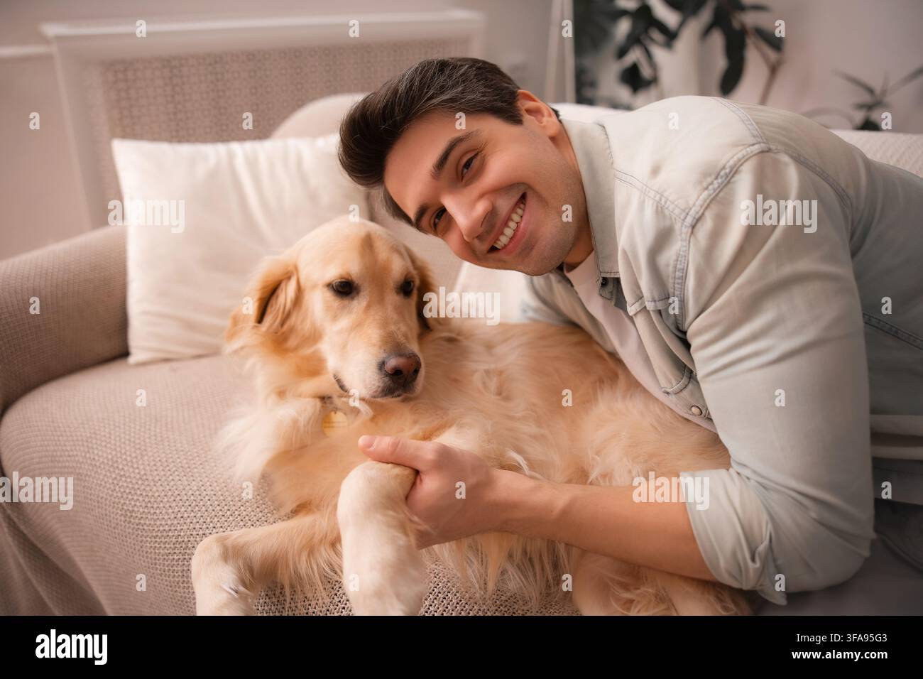 Giovane uomo felice abbraccia un Golden Retriever calmo e ne tiene la zampa mentre si rilassa insieme su un divano a casa. Foto Stock