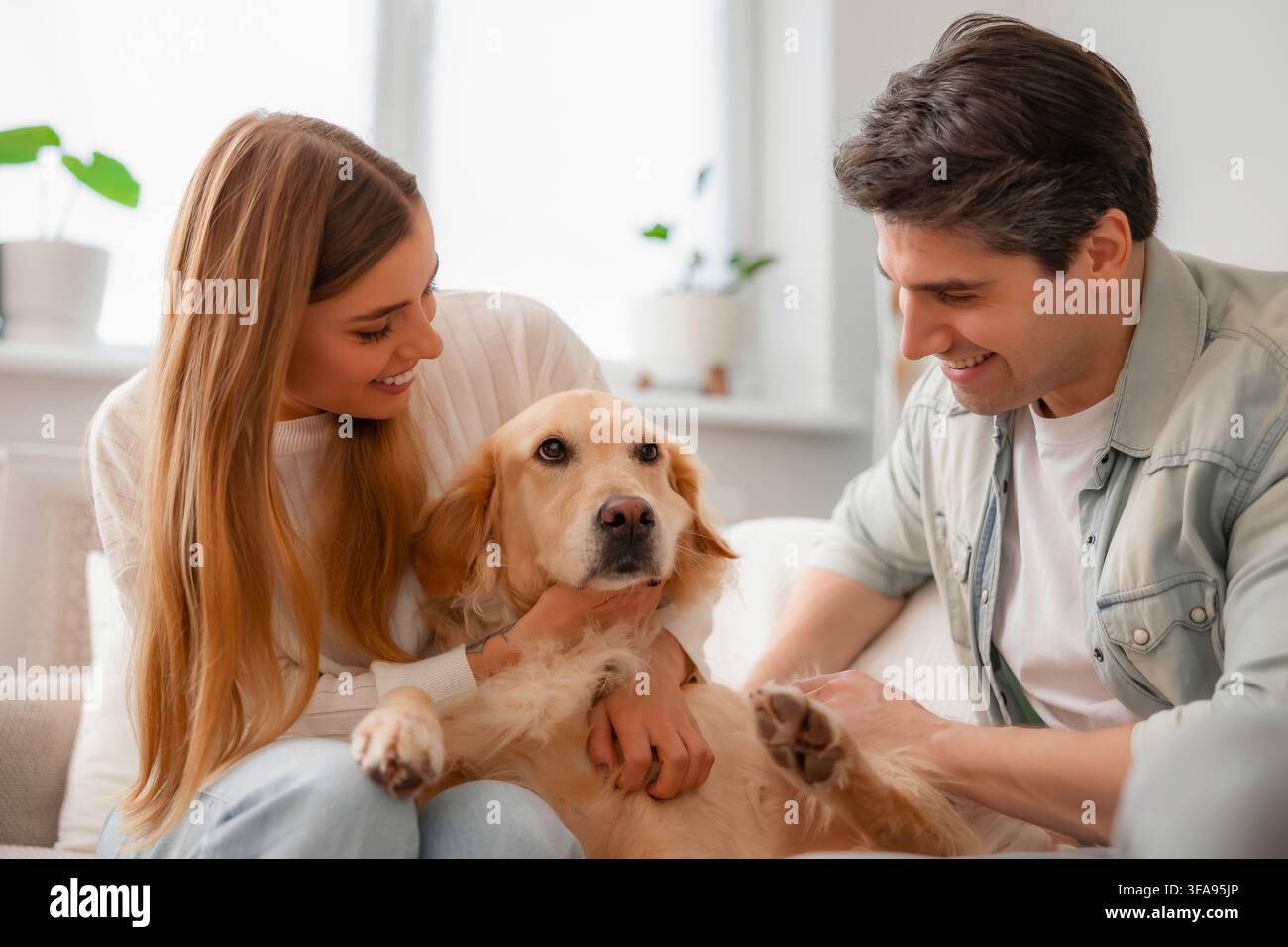 Una giovane coppia affettuosa sorride al loro Golden Retriever mentre coccolano insieme il cane tranquillo su un divano a casa. Foto Stock