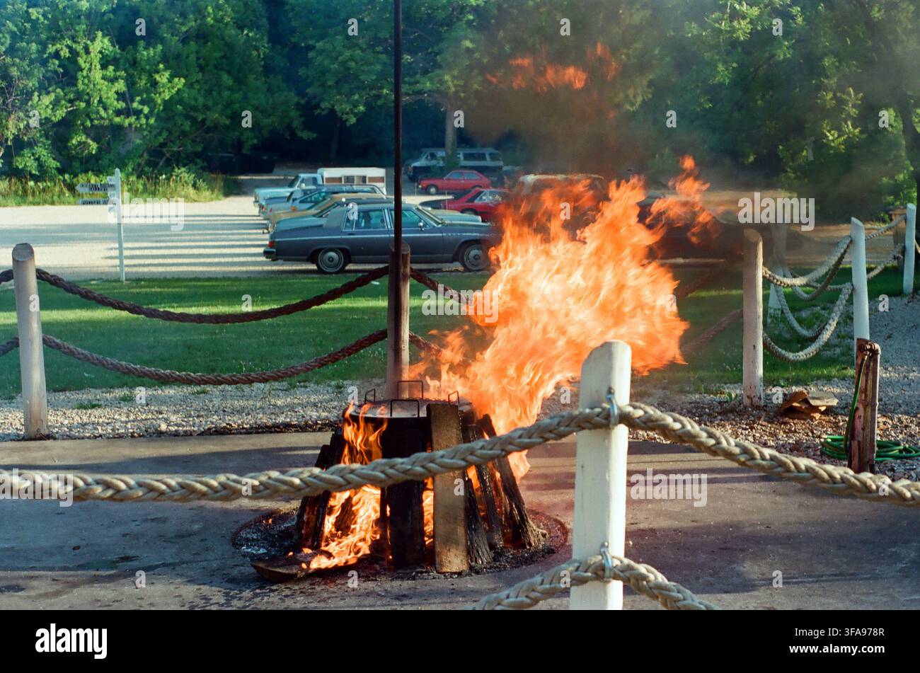 Un grande falò arde intensamente all'interno di una barriera di corde e pali vicino a un parcheggio con auto d'epoca e alberi verdi sullo sfondo. Foto Stock