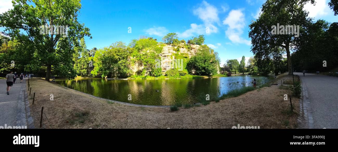 Vista panoramica di un lago sereno del parco circondato da alberi verdi rigogliosi, con una grotta rocciosa sullo sfondo sotto un cielo blu. Foto Stock